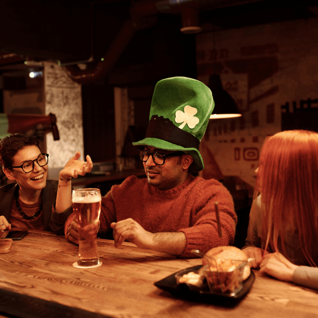Friends celebrating St. Patrick’s Day at a pub, featuring a man wearing a tall green shamrock hat holding a pint of beer while chatting with friends at a wooden bar table.