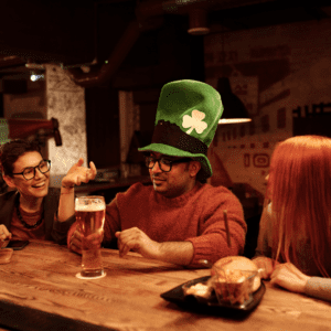 Friends celebrating St. Patrick’s Day at a pub, featuring a man wearing a tall green shamrock hat holding a pint of beer while chatting with friends at a wooden bar table.