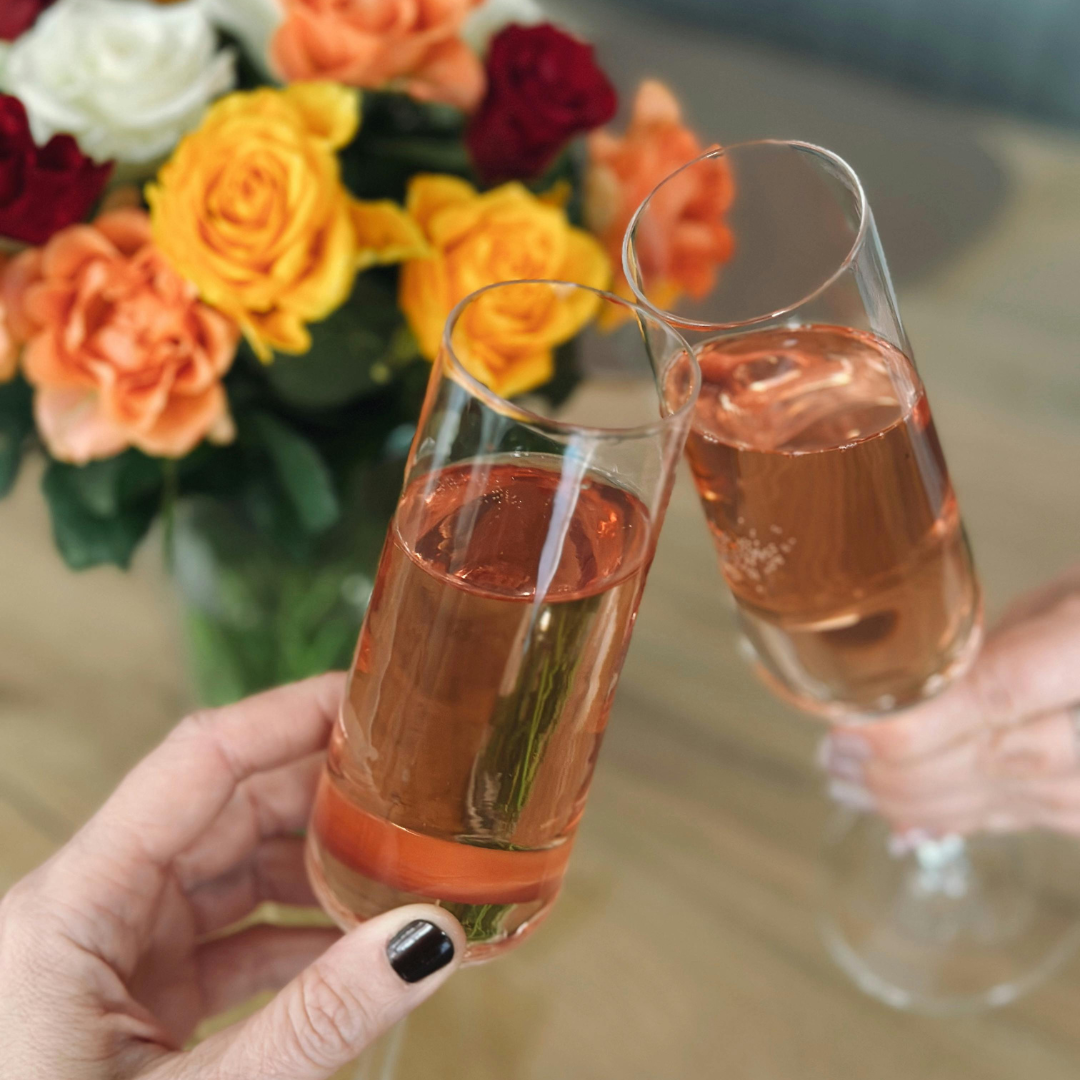 9:58 AMTwo hands clinking stemless champagne flutes filled with rosé, with a colorful bouquet of orange, yellow, and red roses in the background.