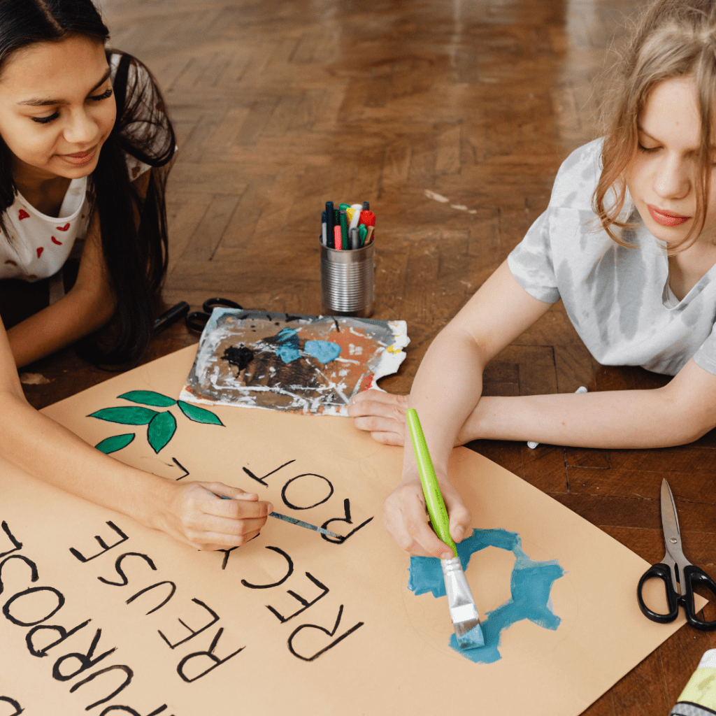 Two young girls paint an environmental awareness poster reading 'Repurpose, Reuse, Recycle' — representing art classes for all ages available this week.