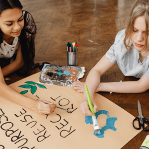 Two young girls paint an environmental awareness poster reading 'Repurpose, Reuse, Recycle' — representing art classes for all ages available this week.