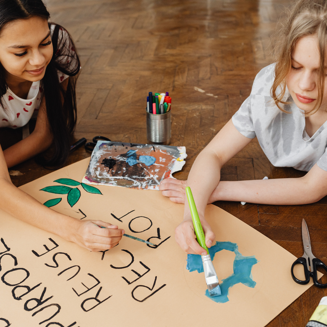 Two young girls paint an environmental awareness poster reading 'Repurpose, Reuse, Recycle' — representing art classes for all ages available this week.