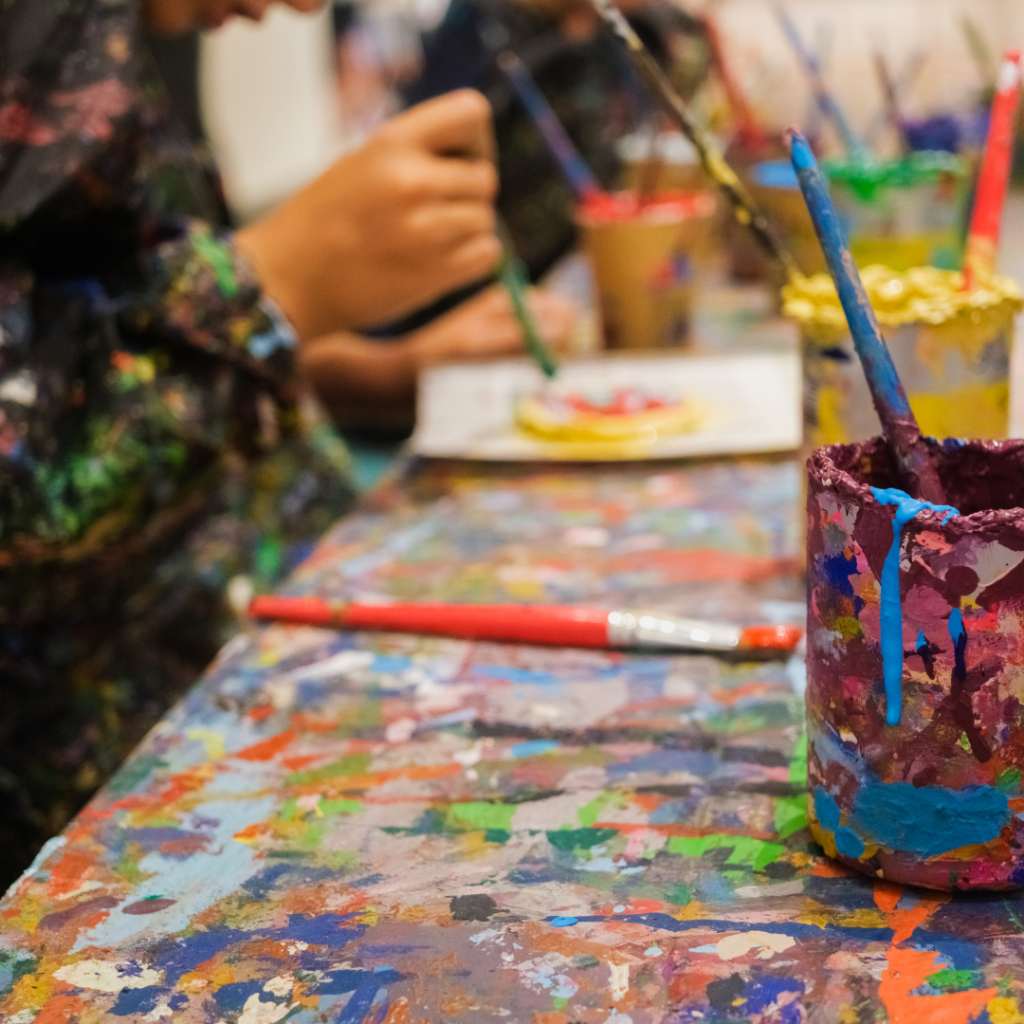 Children and adults participate in a colorful art studio activity, painting and creating artwork at a paint-splattered table filled with brushes and vibrant paint containers