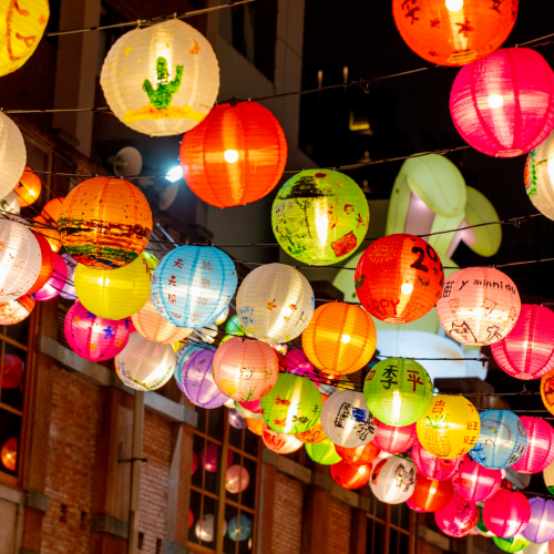 Colorful paper lanterns strung overhead at night, glowing in red, orange, pink, green, blue, and yellow, with hand-drawn designs and characters on them, hanging above a brick-lined street.