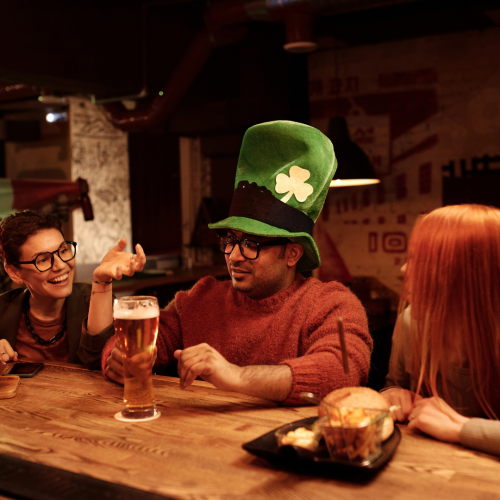 Friends celebrating St. Patrick’s Day at a pub, featuring a man wearing a tall green shamrock hat holding a pint of beer while chatting with friends at a wooden bar table.